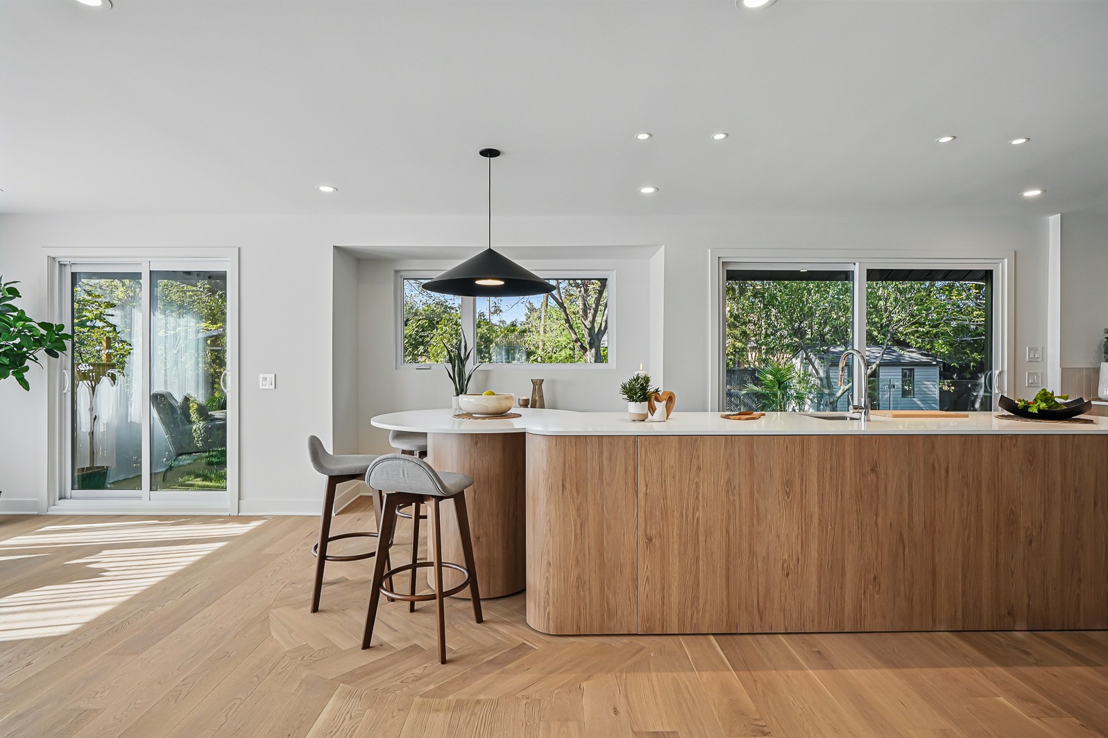 Grande cuisine ouverte avec îlot arrondi en bois, comptoir blanc, suspension noire et plancher de chêne à chevrons.