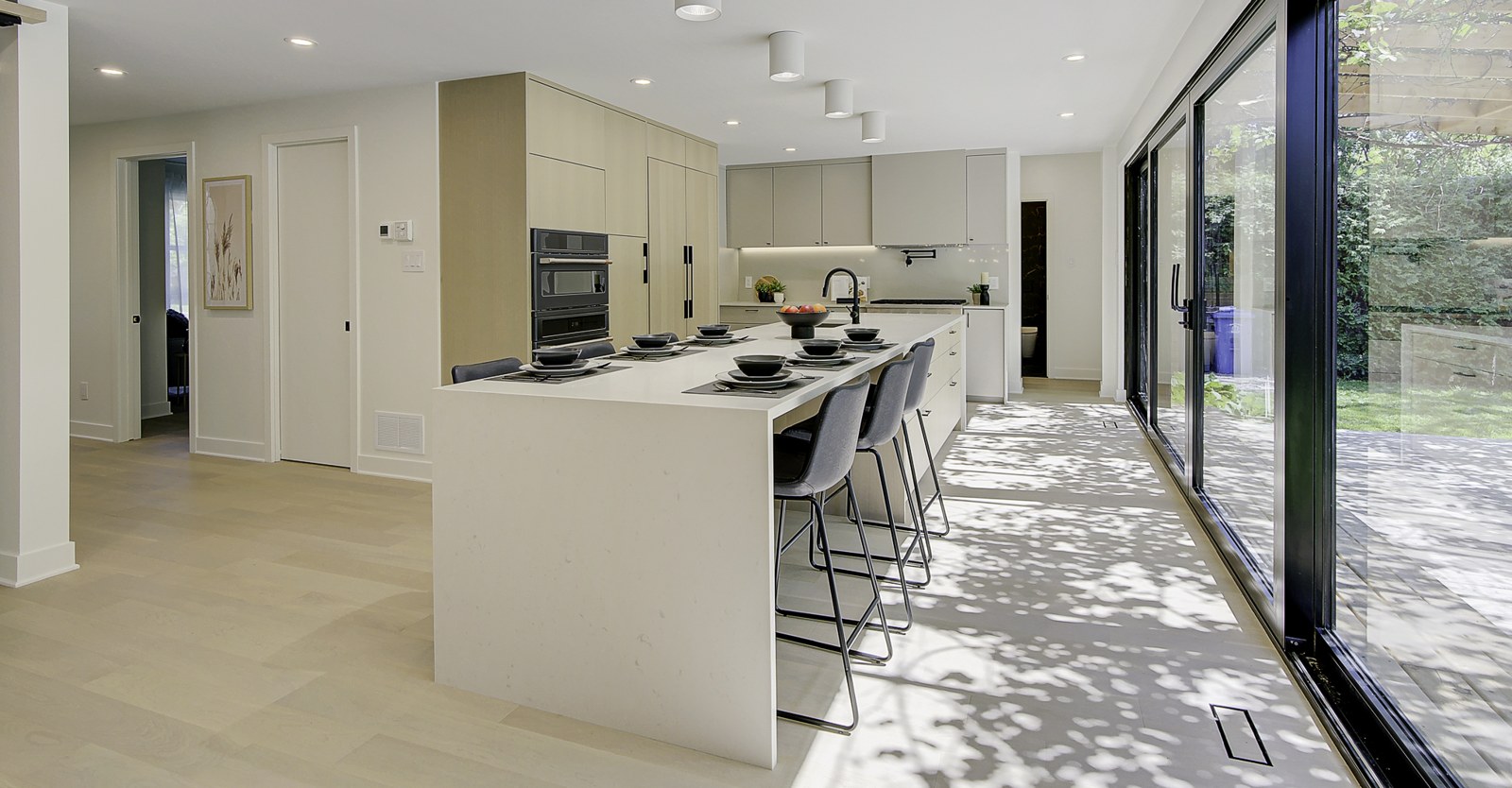 Cuisine ouverte avec long îlot de quartz blanc en cascade, tabourets gris et grande baie vitrée sur la cour.