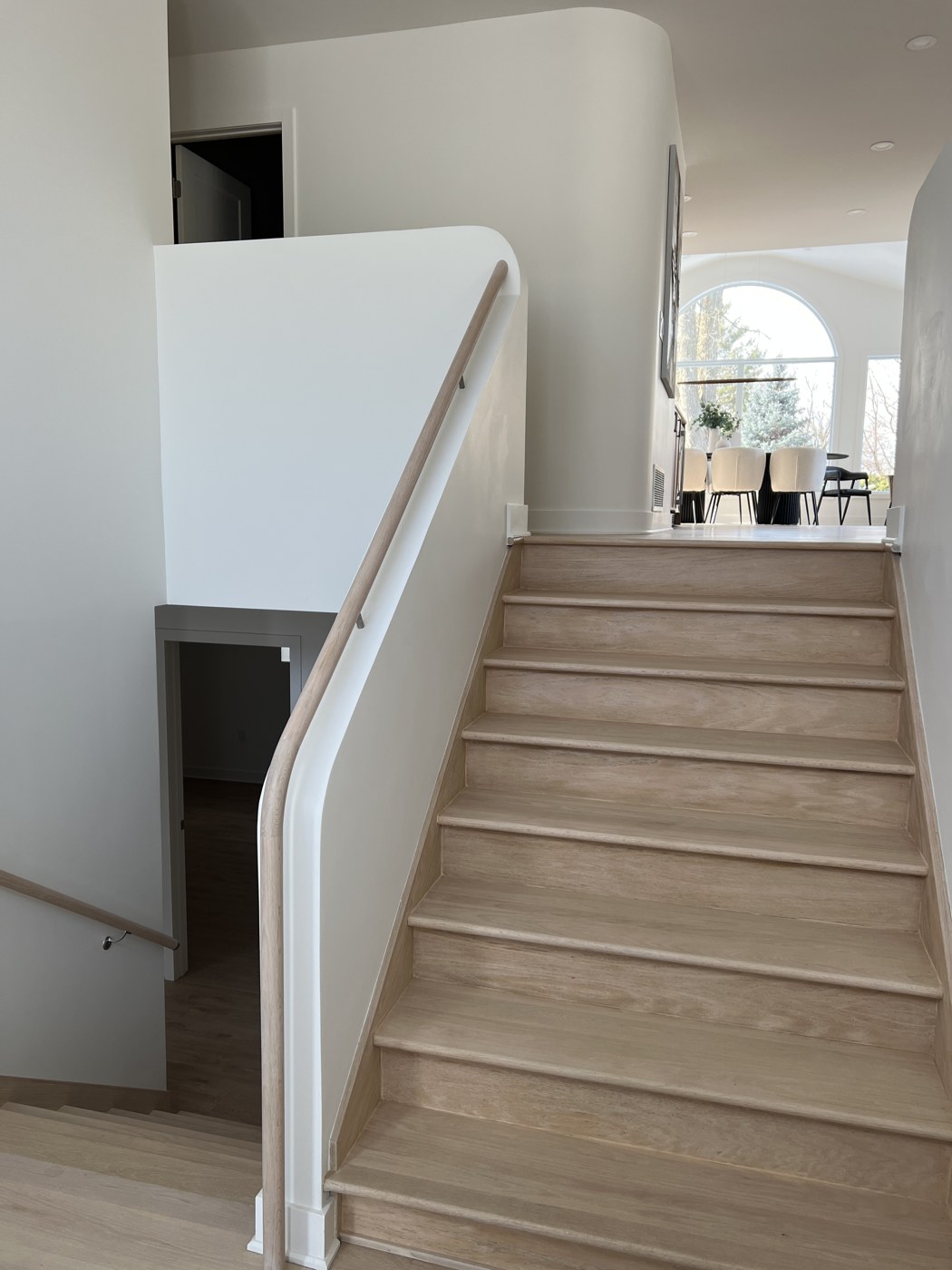 Après : escalier de chêne pâle, murs blancs aux courbes douces et main courante en bois, avec vue sur la salle à manger.